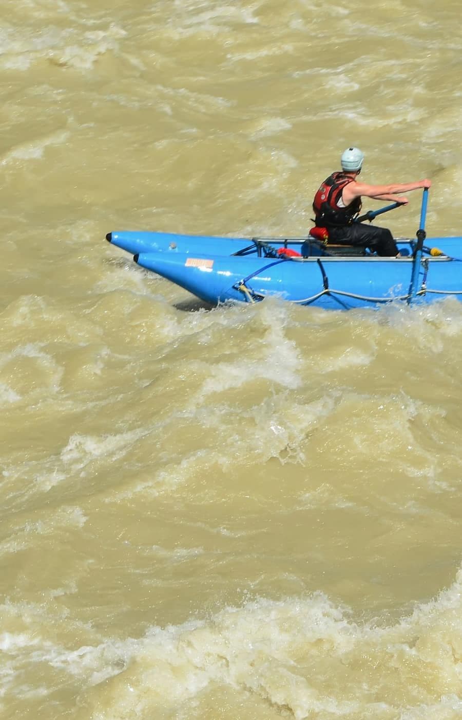 A paddler in a helmet steers a blue raft through muddy whitewater.