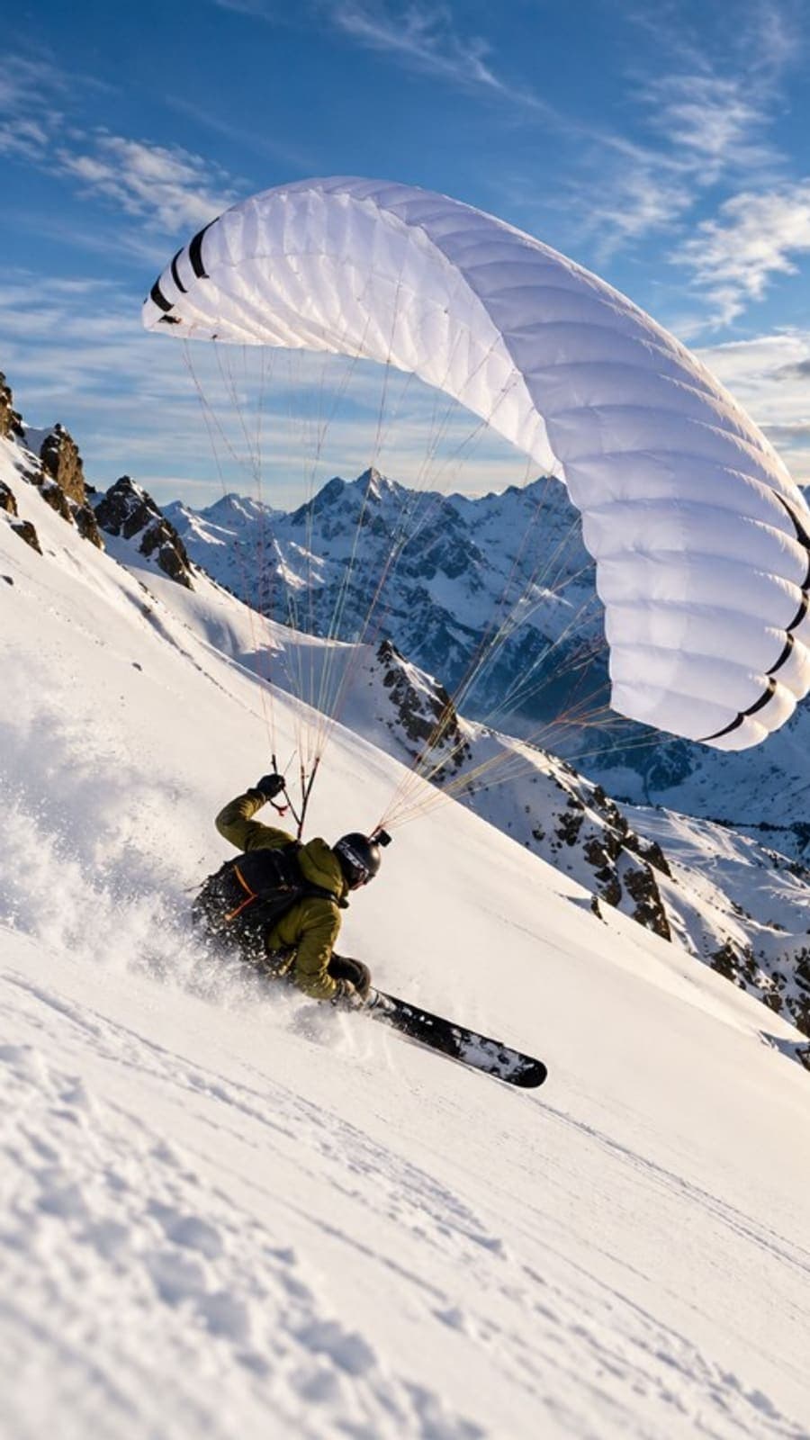 A speedrider in an olive jacket skis a steep face under a white wing, with sun over the peaks.